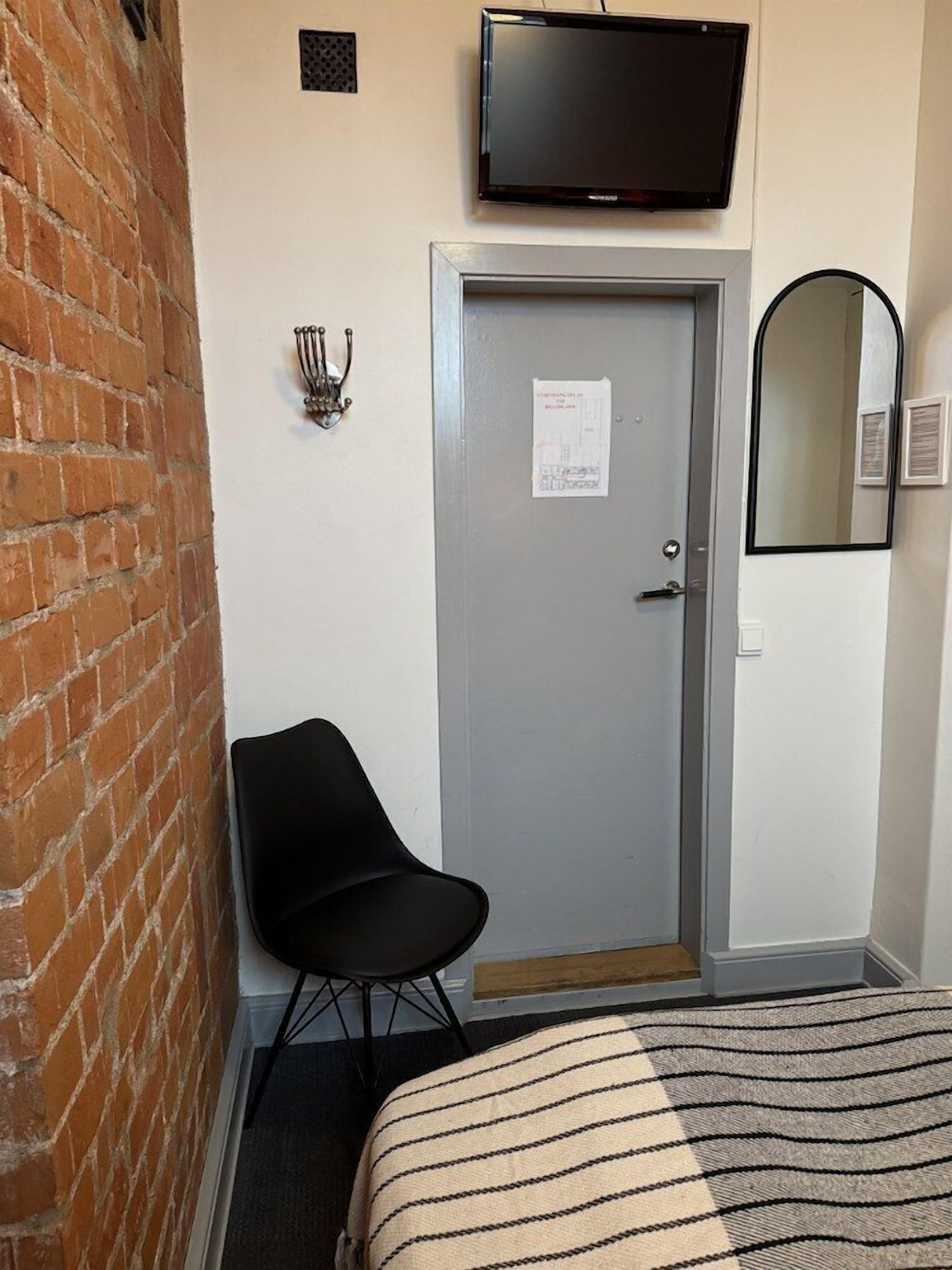 Une chambre d’hôtel avec un mur en briques, une porte grise, une chaise noire, un téléviseur fixé au-dessus de la porte et un miroir sur le mur.