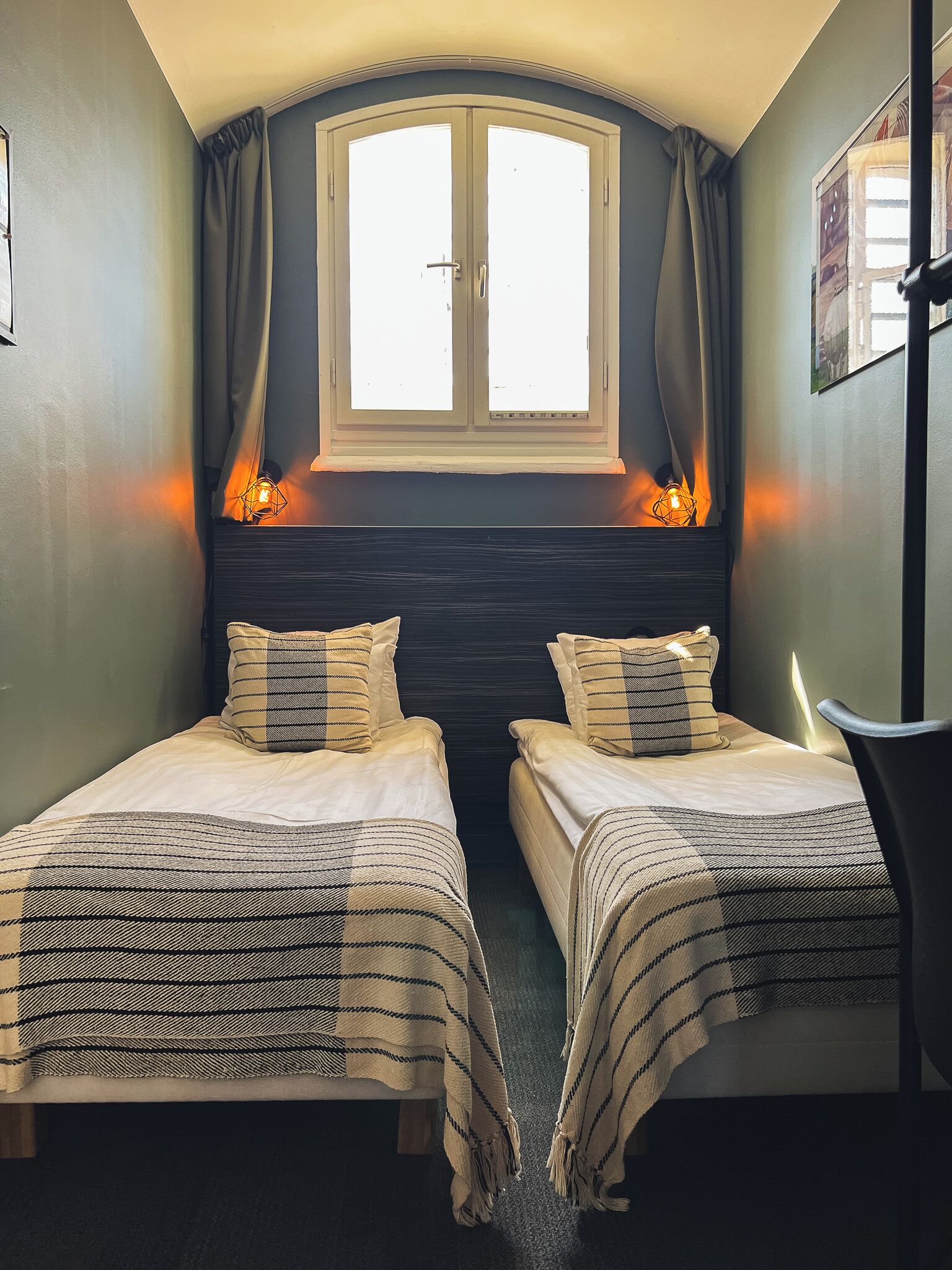 Une chambre avec deux lits, une fenêtre avec des rideaux et un luminaire fixé au mur.