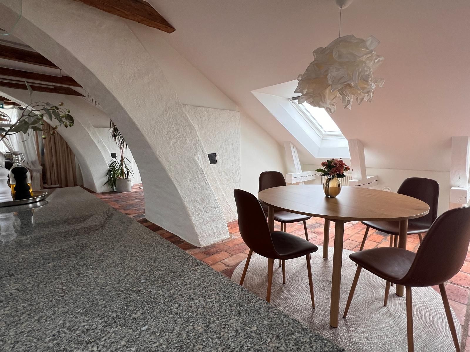 A dining area with a round table, chairs, a hanging light fixture, and a view of the sky through a skylight.