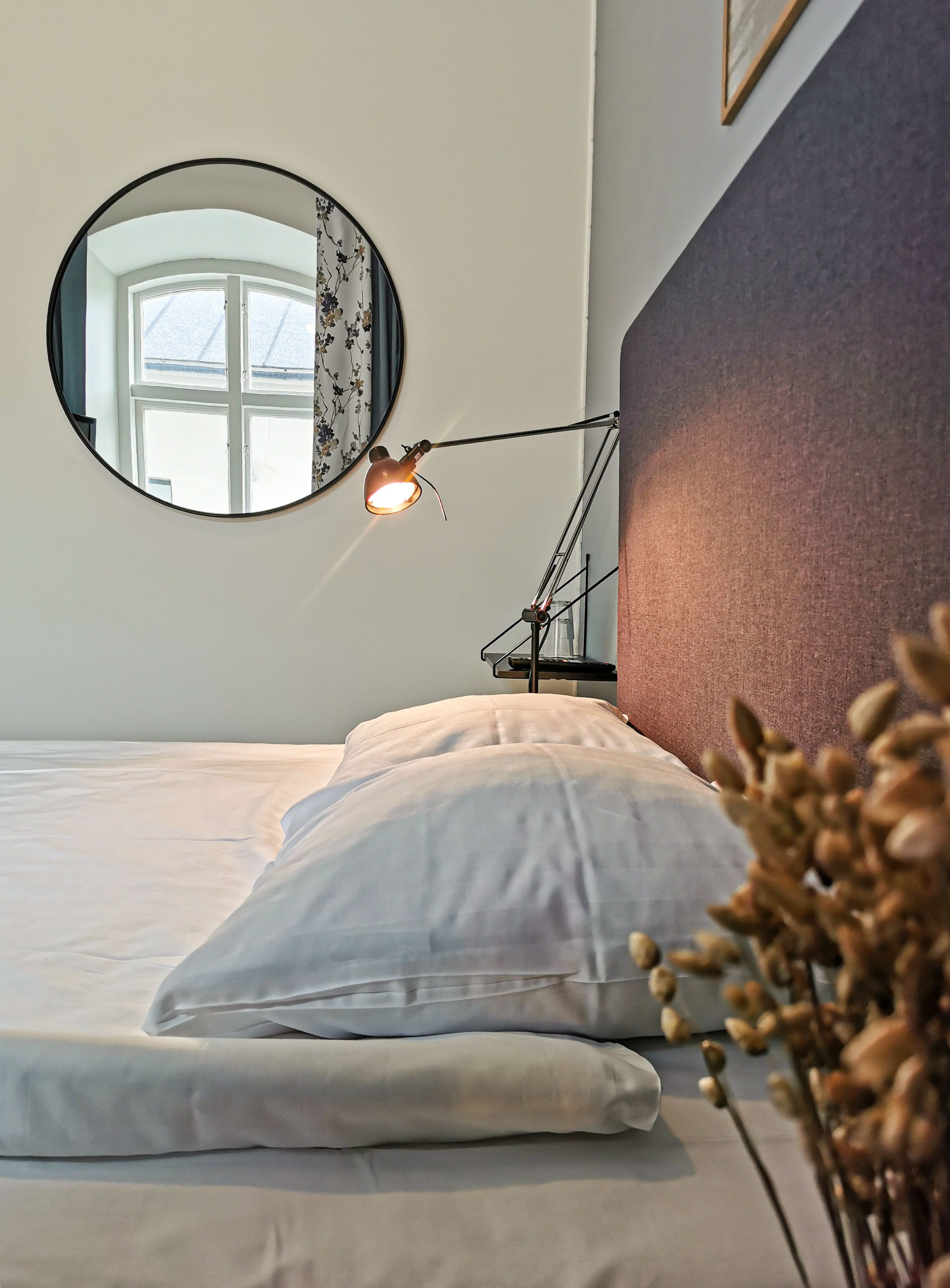 Un lit bien fait, avec du linge de lit blanc, un miroir rond accroché au mur et une lampe de chevet.