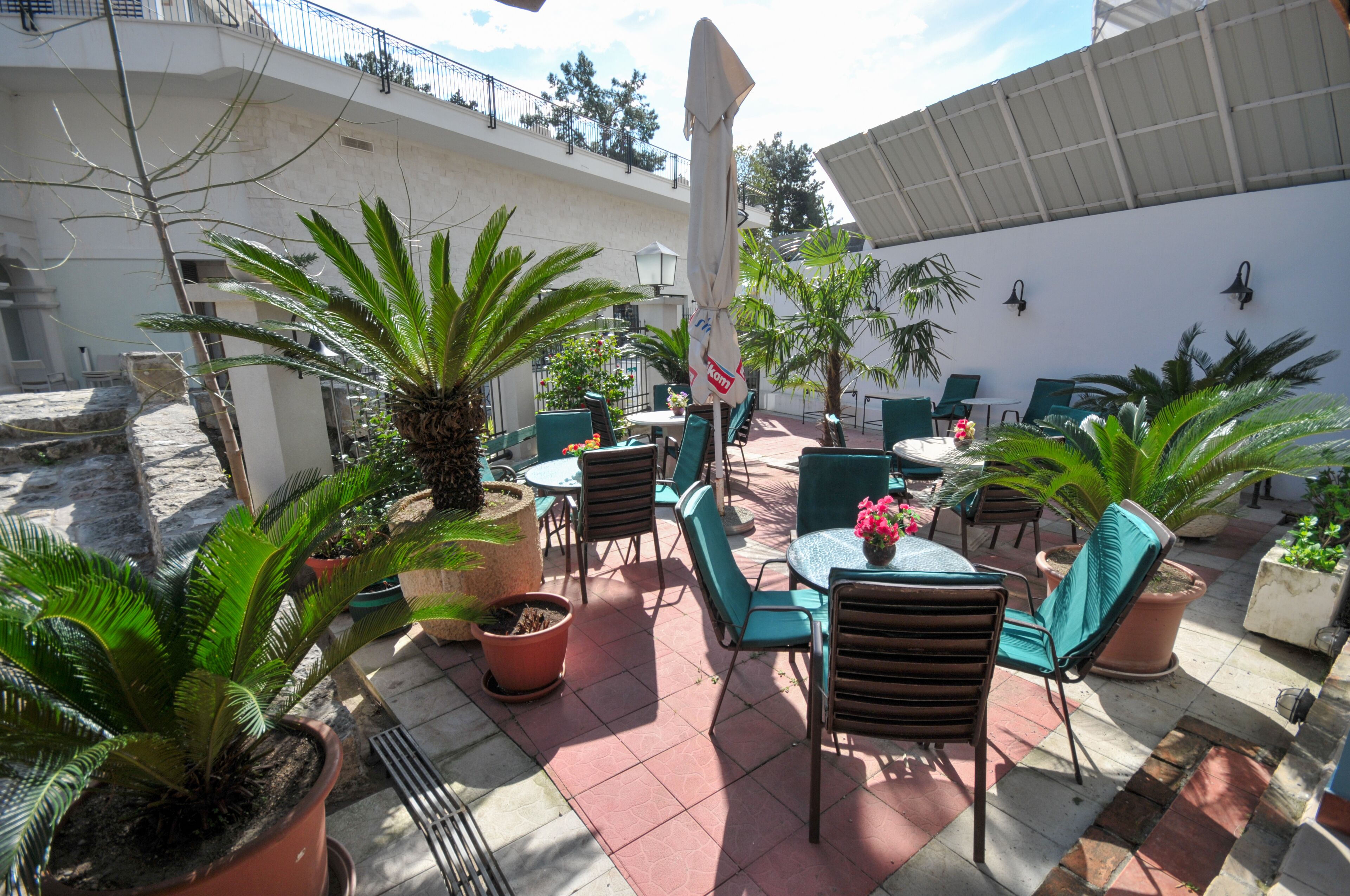 An outdoor patio area with multiple tables and chairs, surrounded by potted plants and a white building in the background.