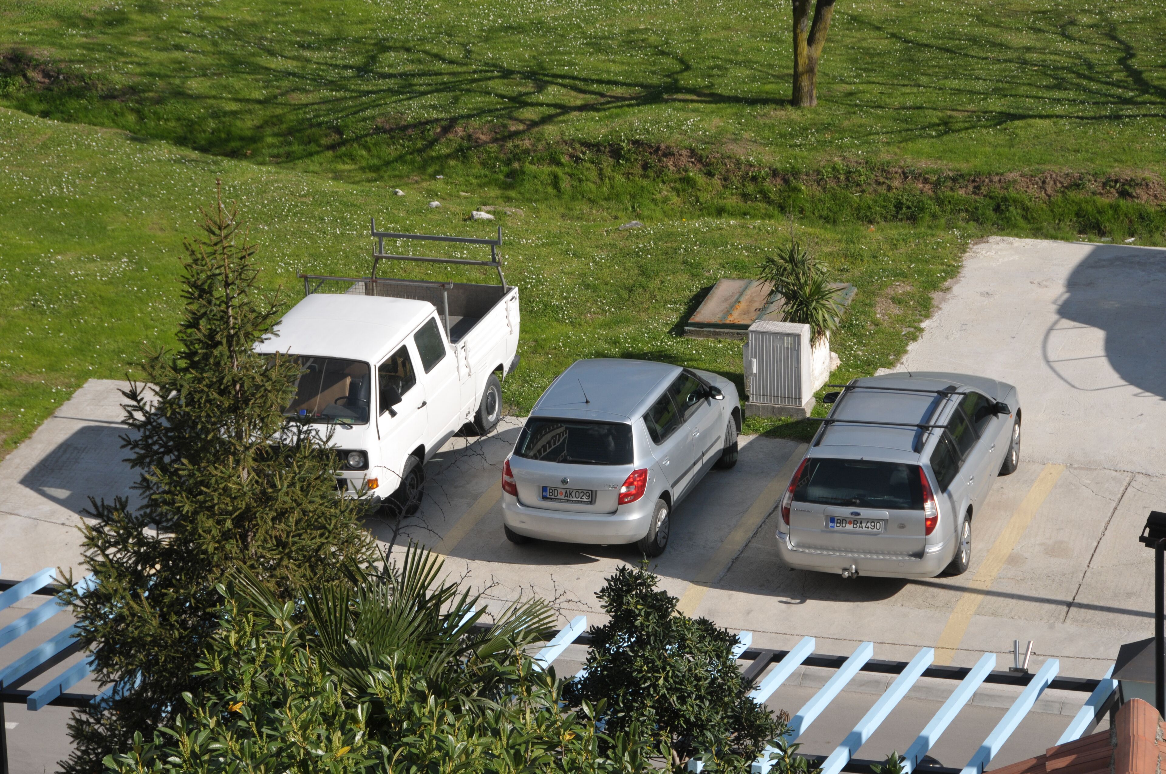 A parking area with three vehicles, a white van, and two silver cars, surrounded by greenery and a clear sky.
