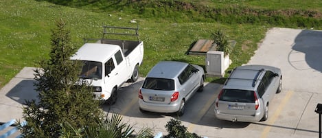 Un estacionamiento con tres vehículos: una camioneta blanca y dos coches plateados, rodeado de vegetación y un cielo despejado.