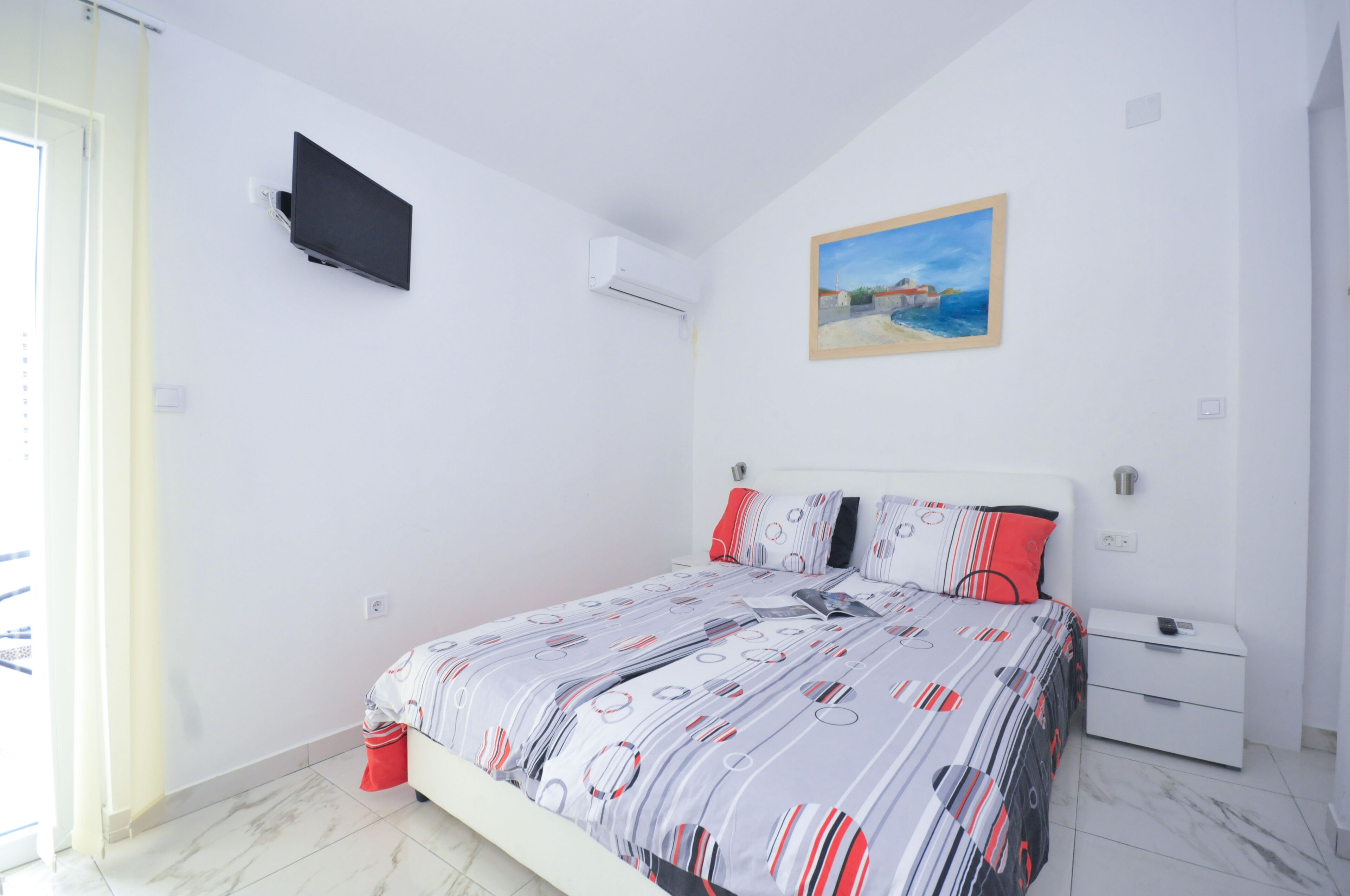 A bedroom with a bed, a TV mounted on the wall, a painting, and a nightstand.