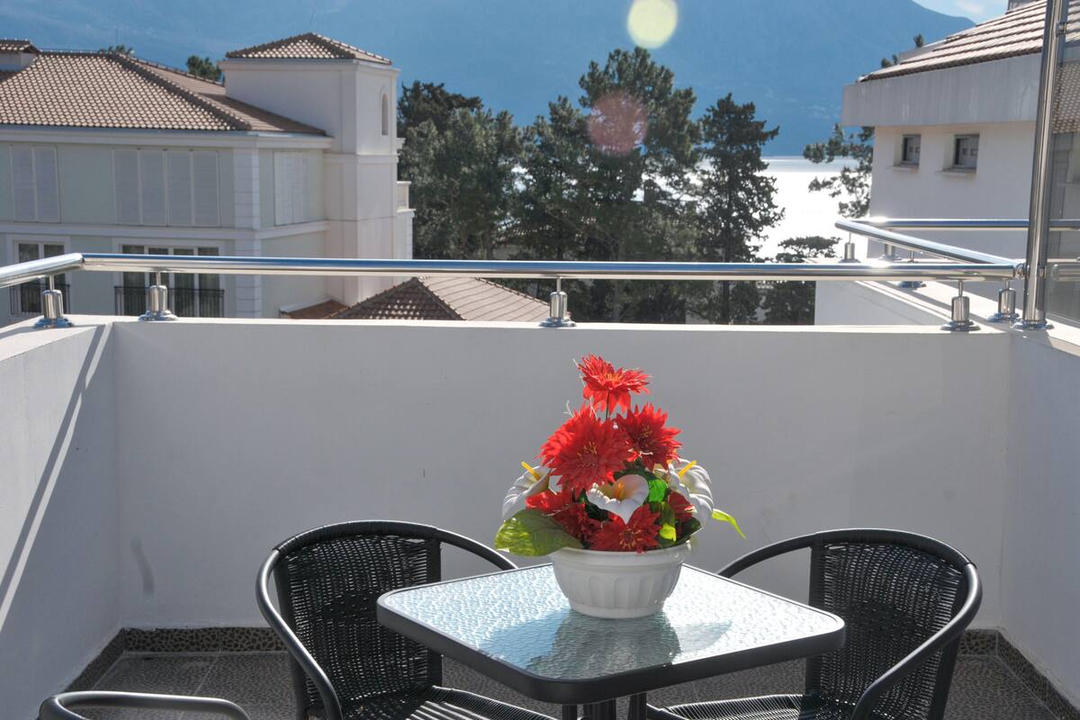 Un balcon avec une table et deux chaises, un pot de fleurs aux fleurs rouges, et une vue dégagée sur les bâtiments et les arbres en arrière-plan.