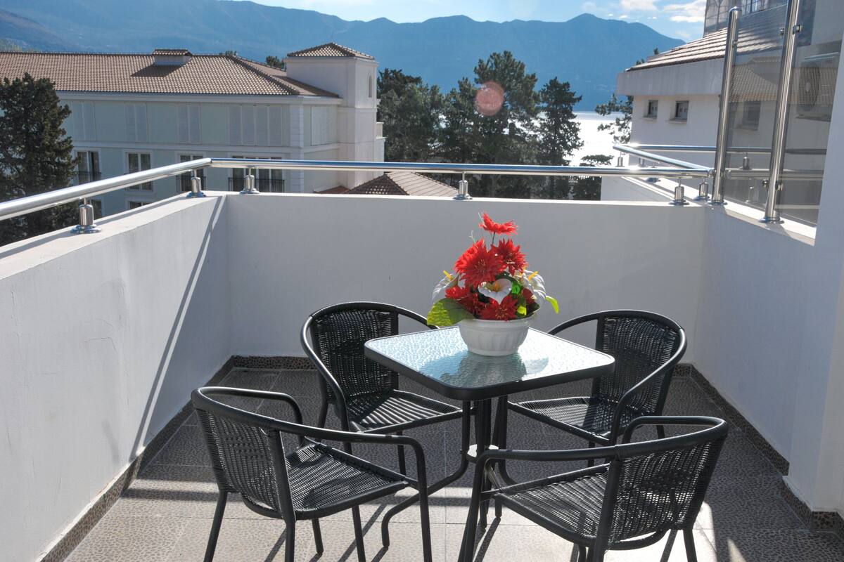 Un balcon avec une table et des chaises, une composition florale et une vue sur des immeubles et des montagnes.