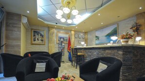 A hotel lobby with a reception desk, black upholstered chairs, a fruit basket, and a painting of a coastal scene. - Oaza (Budva)