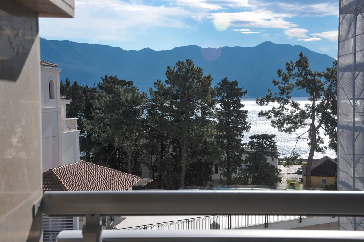 Une vue depuis un balcon donnant sur un lac, avec des montagnes en arrière-plan, des arbres au premier plan et un bâtiment surmonté d’un toit en tuiles.