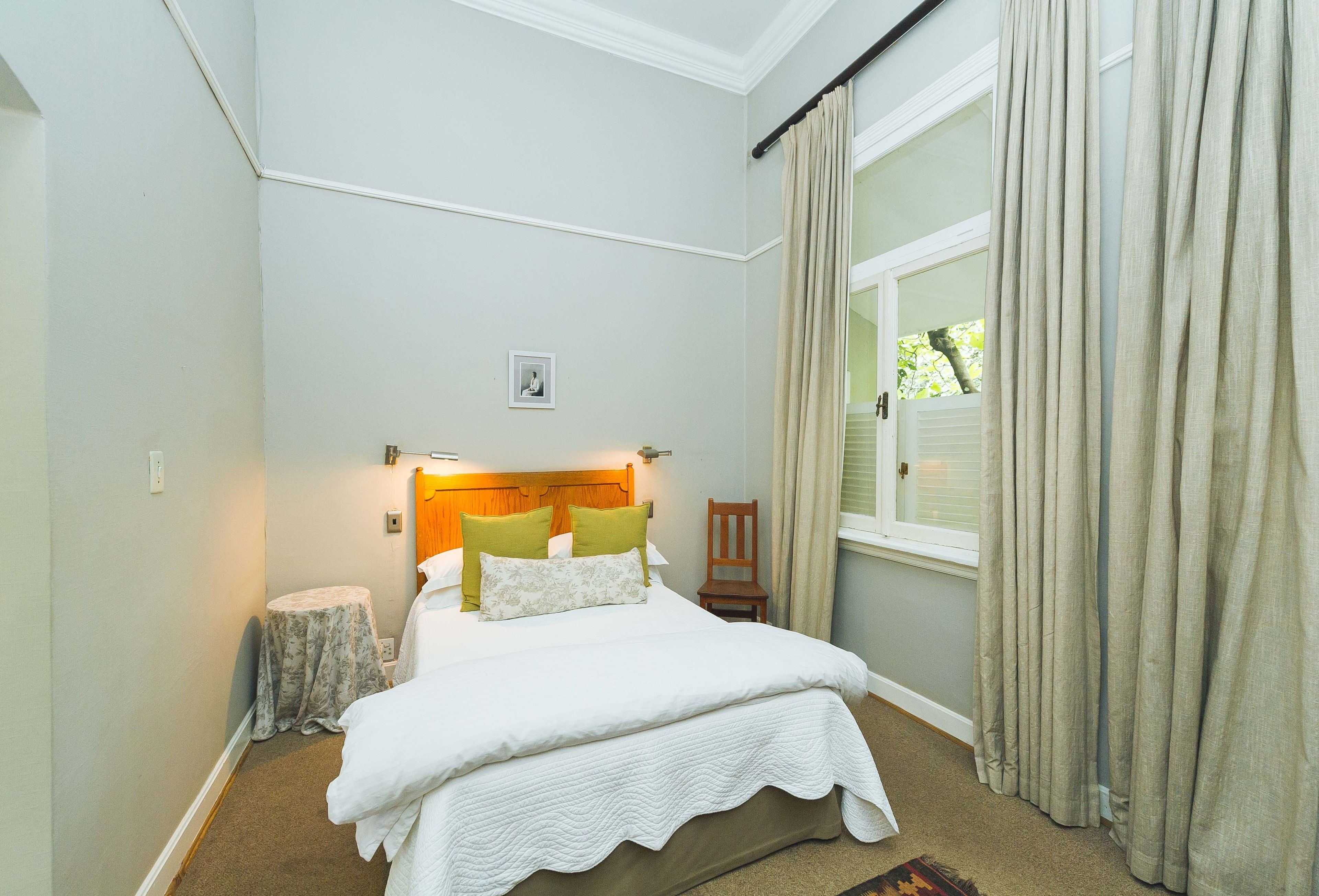 Un dormitorio ordenado con cama, mesita de noche, silla y ventana con cortinas.