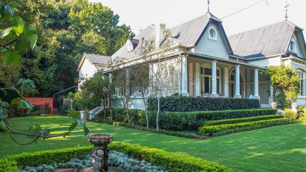 Una casa grande y elegante con un jardín cuidado, que incluye una fuente y setos podados.