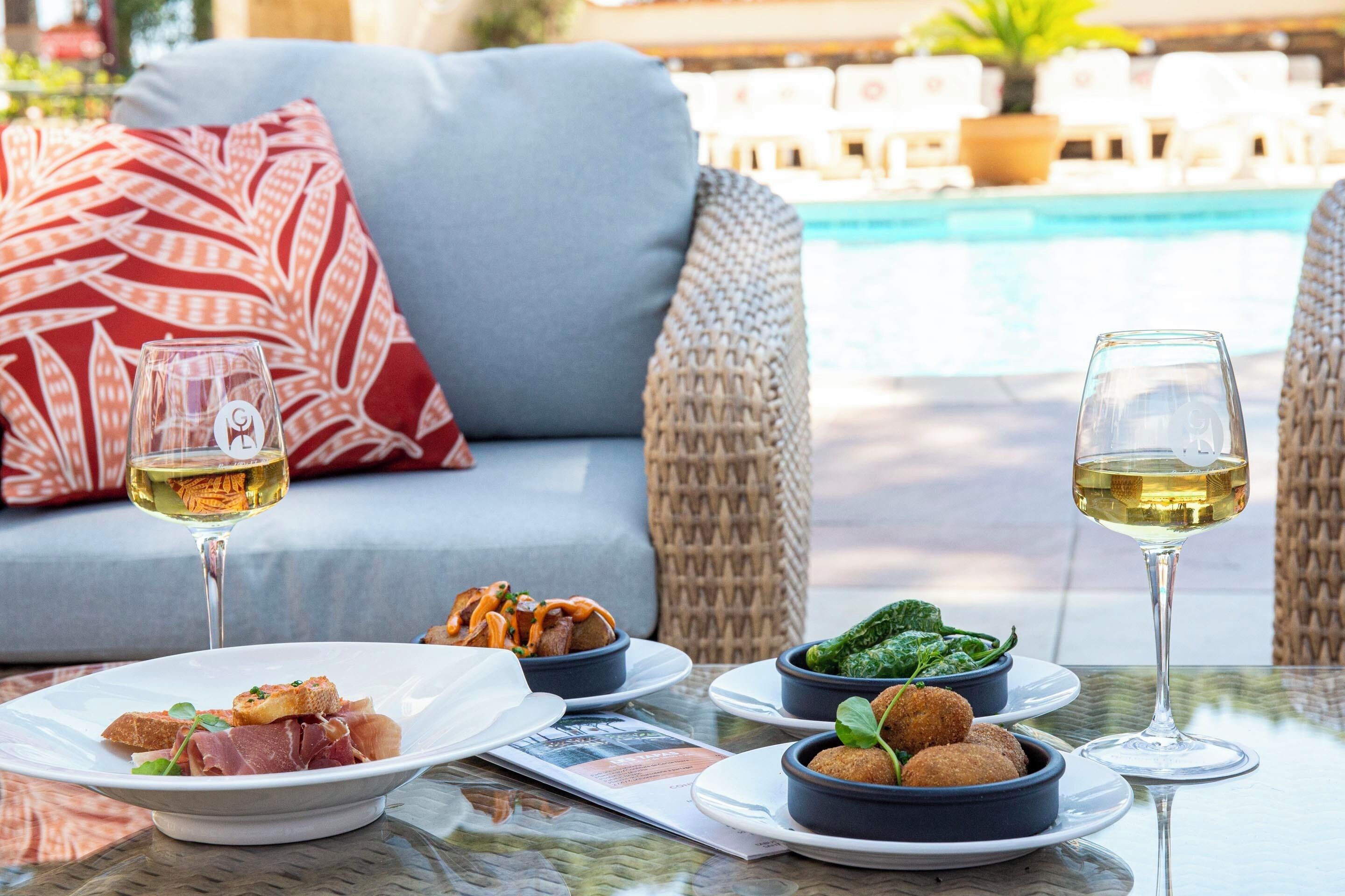 Una mesa de comedor con platos de comida, dos copas de vino blanco y una silla de mimbre con un cojín rojo al fondo.