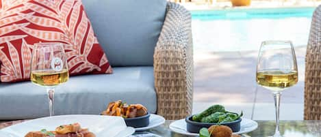 A dining table with plates of food, two glasses of white wine, and a wicker chair with a red pillow in the background.
