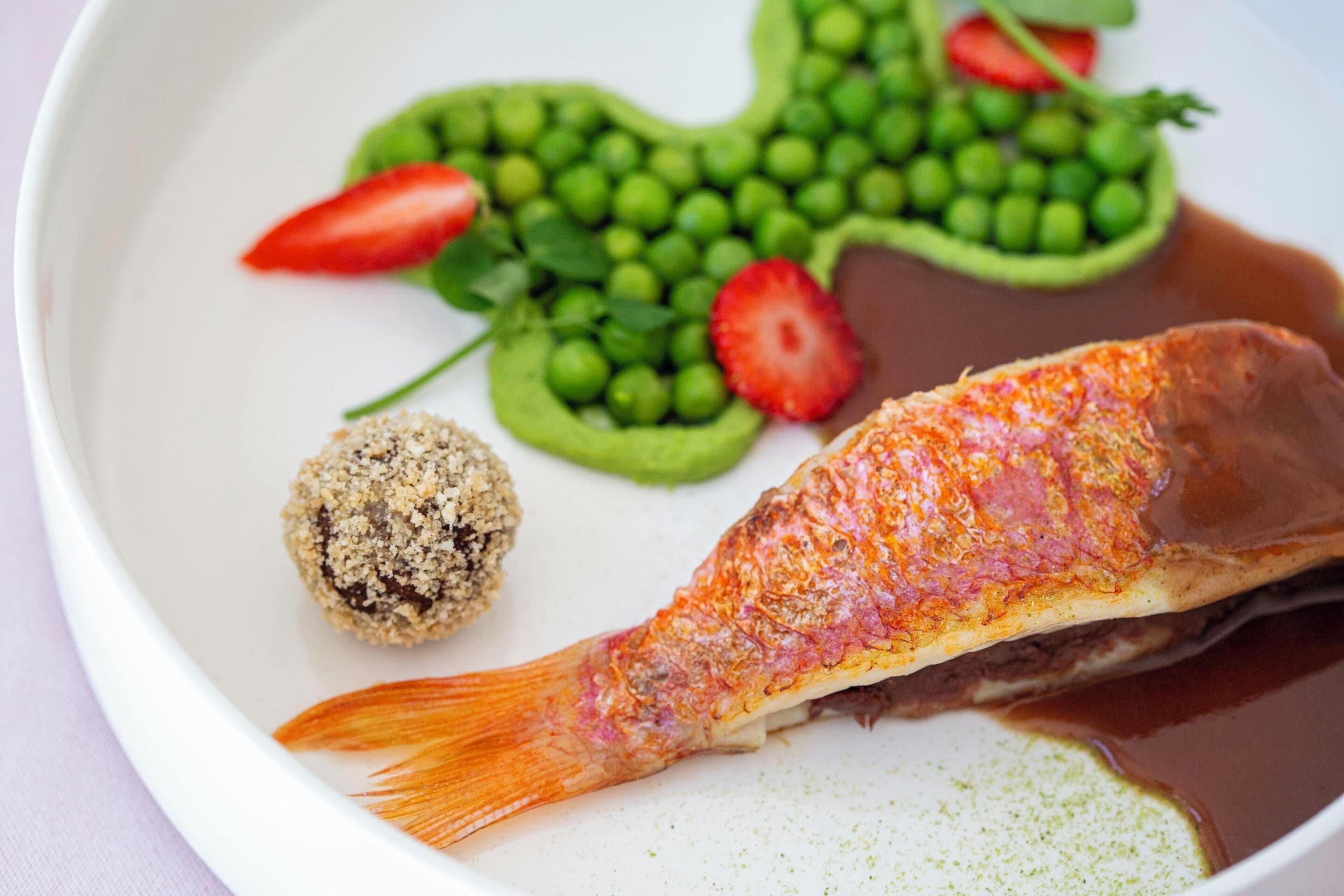A plate with a fish, peas, strawberries, and a round food item.