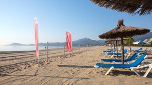 A beach with rows of blue and white lounge chairs, a thatched umbrella, and red flags. - Grand Hôtel du Lido (Argelès-sur-Mer)