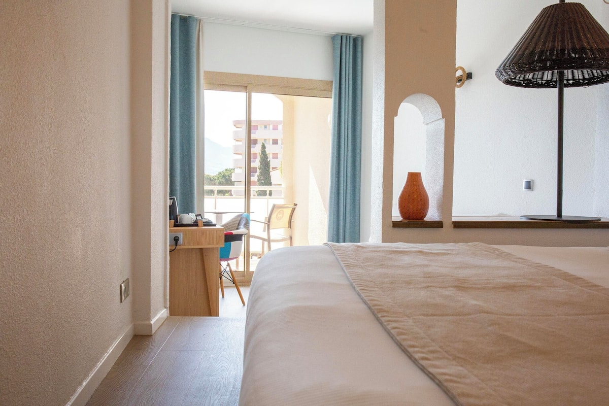 Una habitación de hotel con una cama grande, un escritorio con silla, una ventana con cortinas y una lámpara.