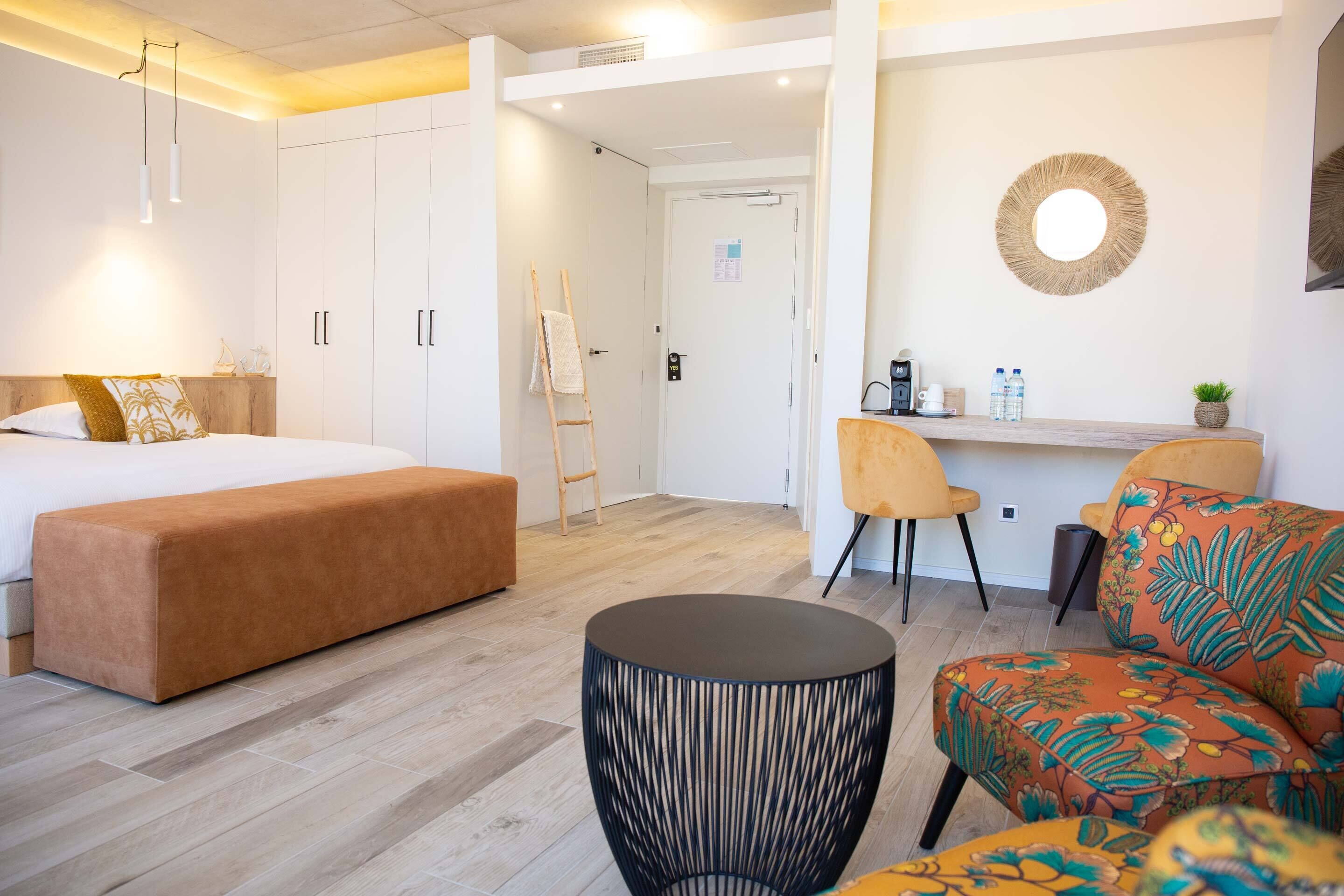 A modern hotel room with a large bed, a round black side table, a sofa with a floral pattern, and a desk with chairs.
