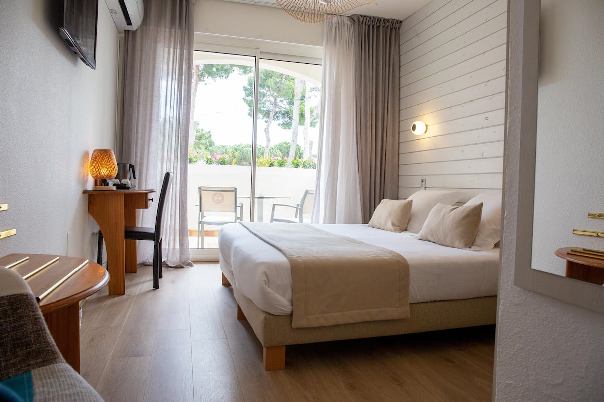 Habitación de hotel con cama, escritorio con lámpara, silla y vistas al exterior a través de una ventana.