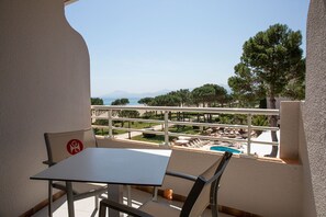 Un balcón con mesa y sillas, con vista a una piscina y un jardín ajardinado con árboles, y una vista distante del mar y las montañas.