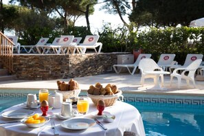 Desayuno junto a la piscina con sillones de descanso, una mesa con alimentos para el desayuno y una pared de piedra al fondo.