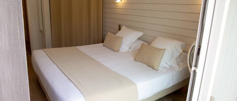 A neatly made bed with a beige blanket and pillows, a wall-mounted light fixture, and wooden paneling on the wall.