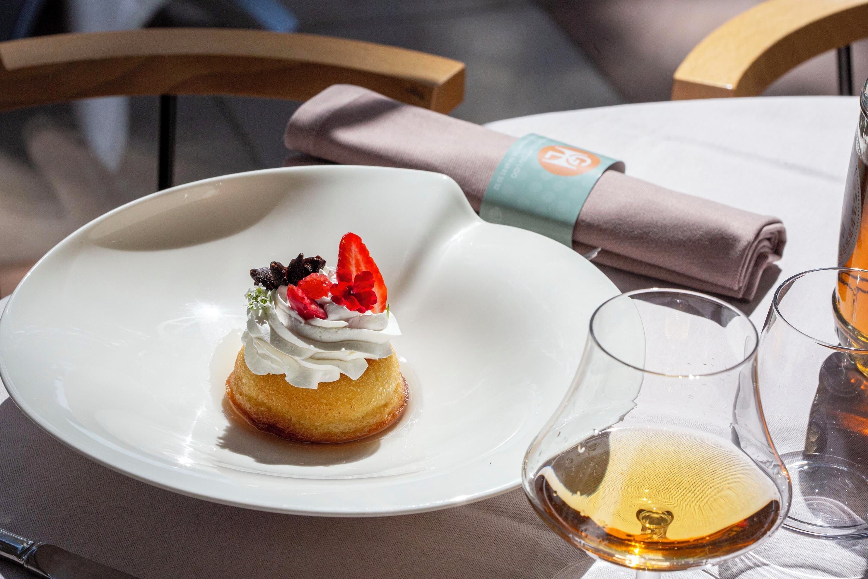A dessert on a white plate with whipped cream and fruit on top, next to a glass of wine on a table.