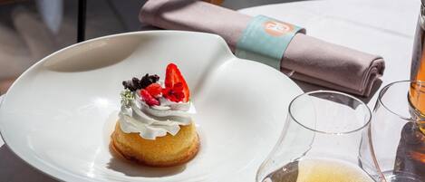 A dessert on a white plate with whipped cream and fruit on top, next to a glass of wine on a table.