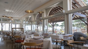 A spacious restaurant with high ceilings, large windows, and modern lighting fixtures. Tables are neatly arranged with chairs, and the view includes outdoor seating and trees. - Grand Hôtel du Lido (Argelès-sur-Mer)