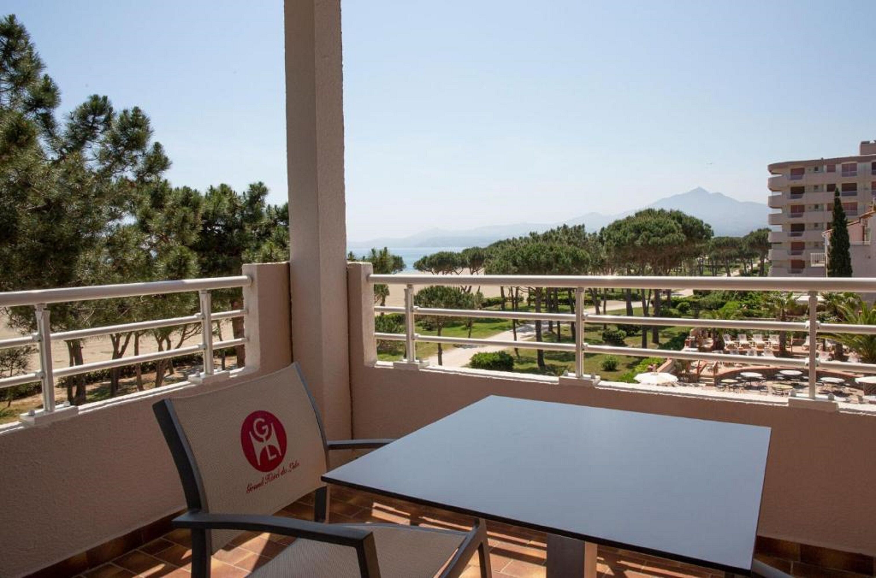 A balcony with a table and chairs, overlooking a landscaped area with trees and a distant mountain.