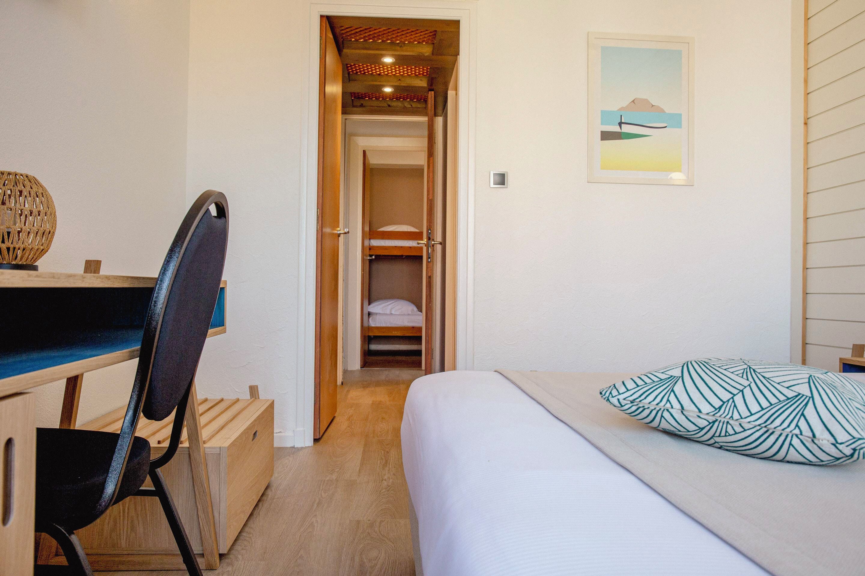 A bedroom with a bed, a desk, and a chair. There is a door leading to a closet with a bunk bed. A picture hangs on the wall.