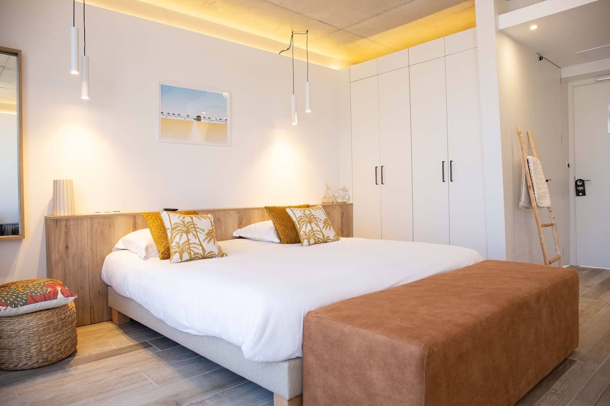 Un dormitorio moderno con una cama grande, cabecera de madera y un banco. Hay un cuadro en la pared y una escalera apoyada en la puerta.