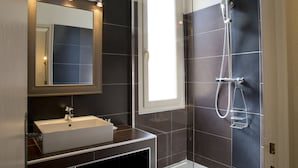 Ein modernes Badezimmer mit großem Spiegel, Waschbecken, Dusche inklusive Handbrause und dunkel gefliesten Wänden.