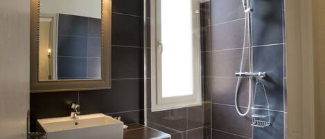 A modern bathroom with a large mirror, a sink, a shower with a handheld showerhead, and dark tiled walls.