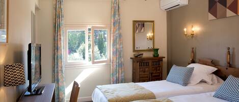 A hotel room with two beds, a desk, a TV, a chandelier, an air conditioner, and a window with curtains.