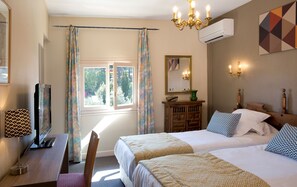 Een hotelkamer met twee bedden, een bureau, een televisie, een kroonluchter, airconditioning en een raam met gordijnen.