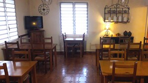Un comedor con mesas y sillas de madera, un televisor sobre un mueble bajo, una ventana con persianas y un estante en la pared con adornos.