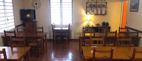A dining area with wooden tables and chairs, a television on a stand, a window with blinds, and a wall-mounted shelf with decorative items.