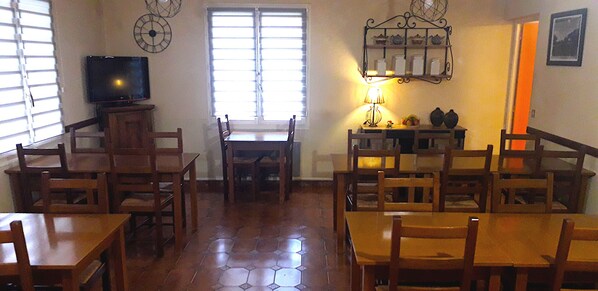A dining area with wooden tables and chairs, a television on a stand, a window with blinds, and a wall-mounted shelf with decorative items. - Hôtel Saint-michel (Digne-les-Bains)
