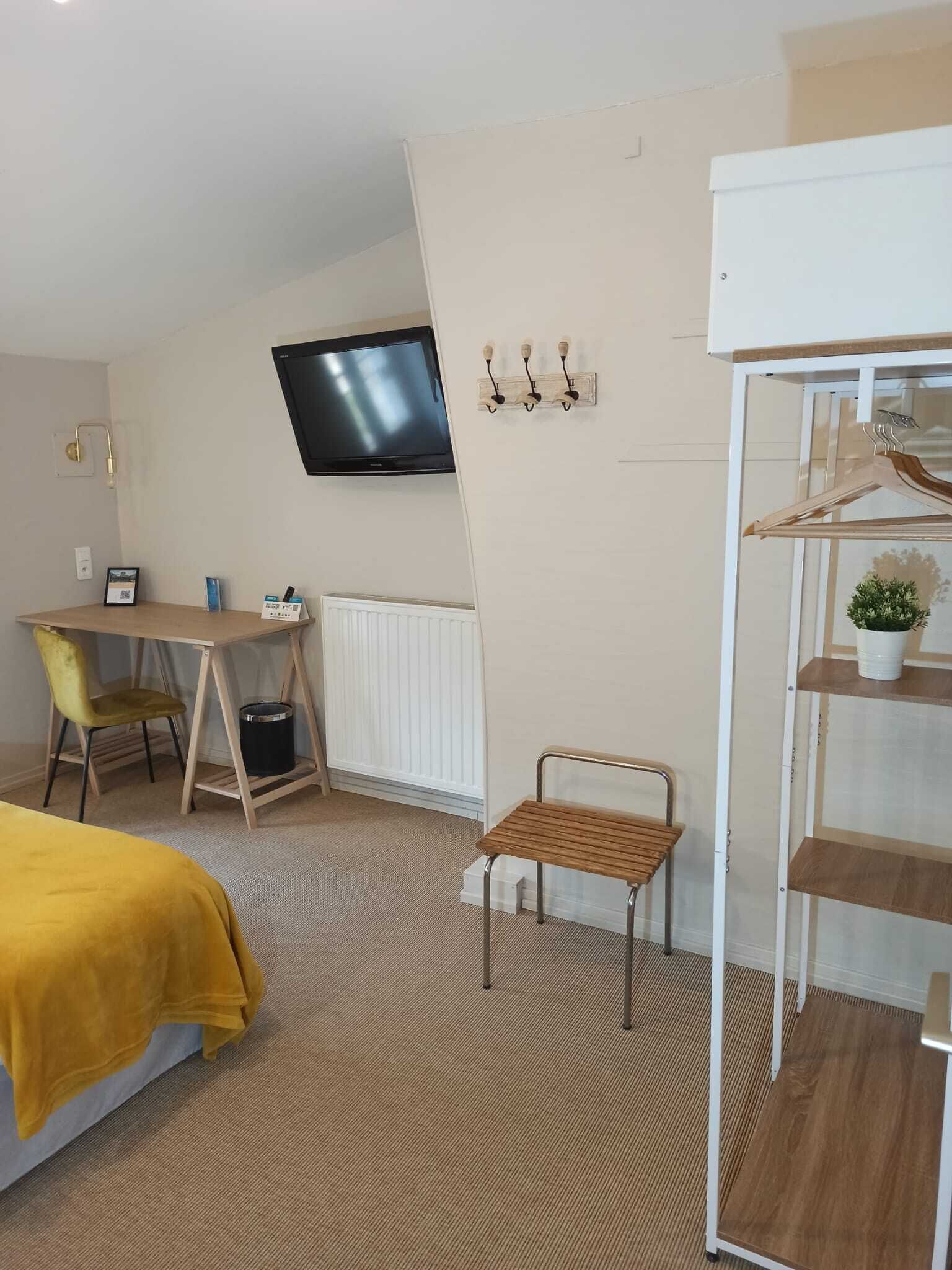 Ein kleines Hotelzimmer mit Bett, Schreibtisch, Stuhl, einem an der Wand befestigten Fernseher und einem kleinen Regal mit einer Pflanze.