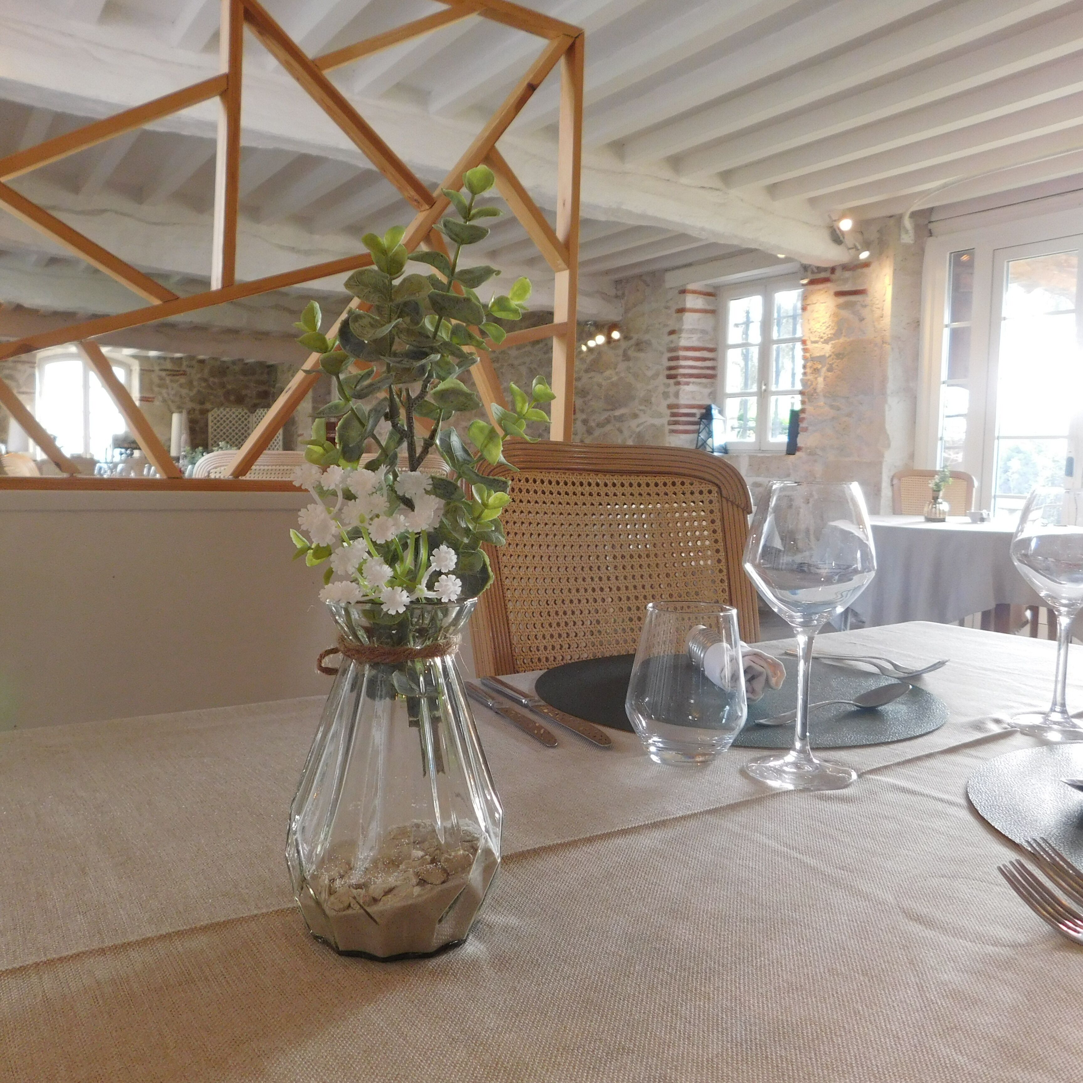 A dining table with a glass vase containing white flowers, a wicker chair, and wine glasses.