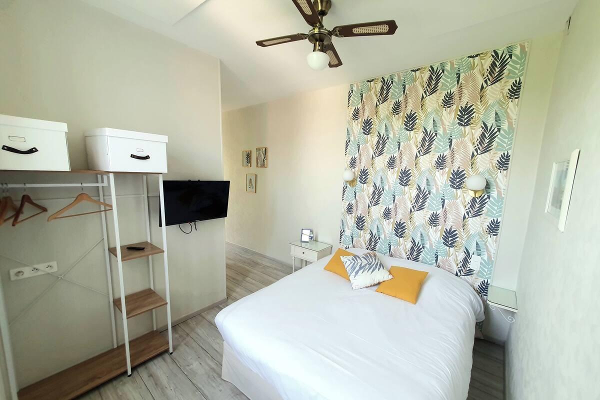 A bedroom with a bed, a wardrobe, a ceiling fan, a TV, and a patterned curtain.