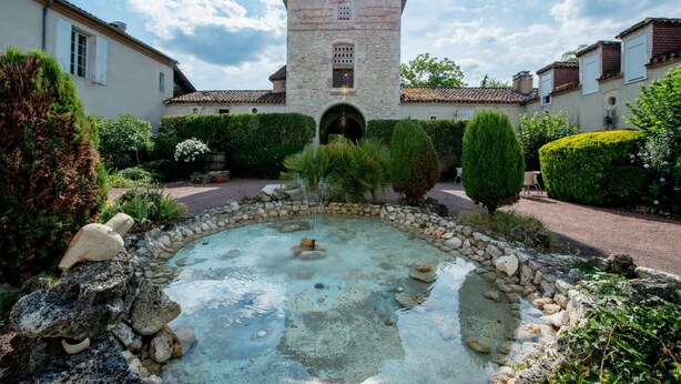Un patio central con una fuente, rodeado de muros de piedra y vegetación, que conduce a un edificio con una torre.