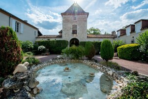 Un patio central con una fuente, rodeado de muros de piedra y vegetación, que conduce a un edificio con una torre.