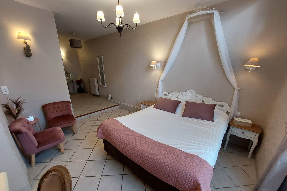 A bedroom with a large bed, a sitting area with a chair, a nightstand, and a chandelier.