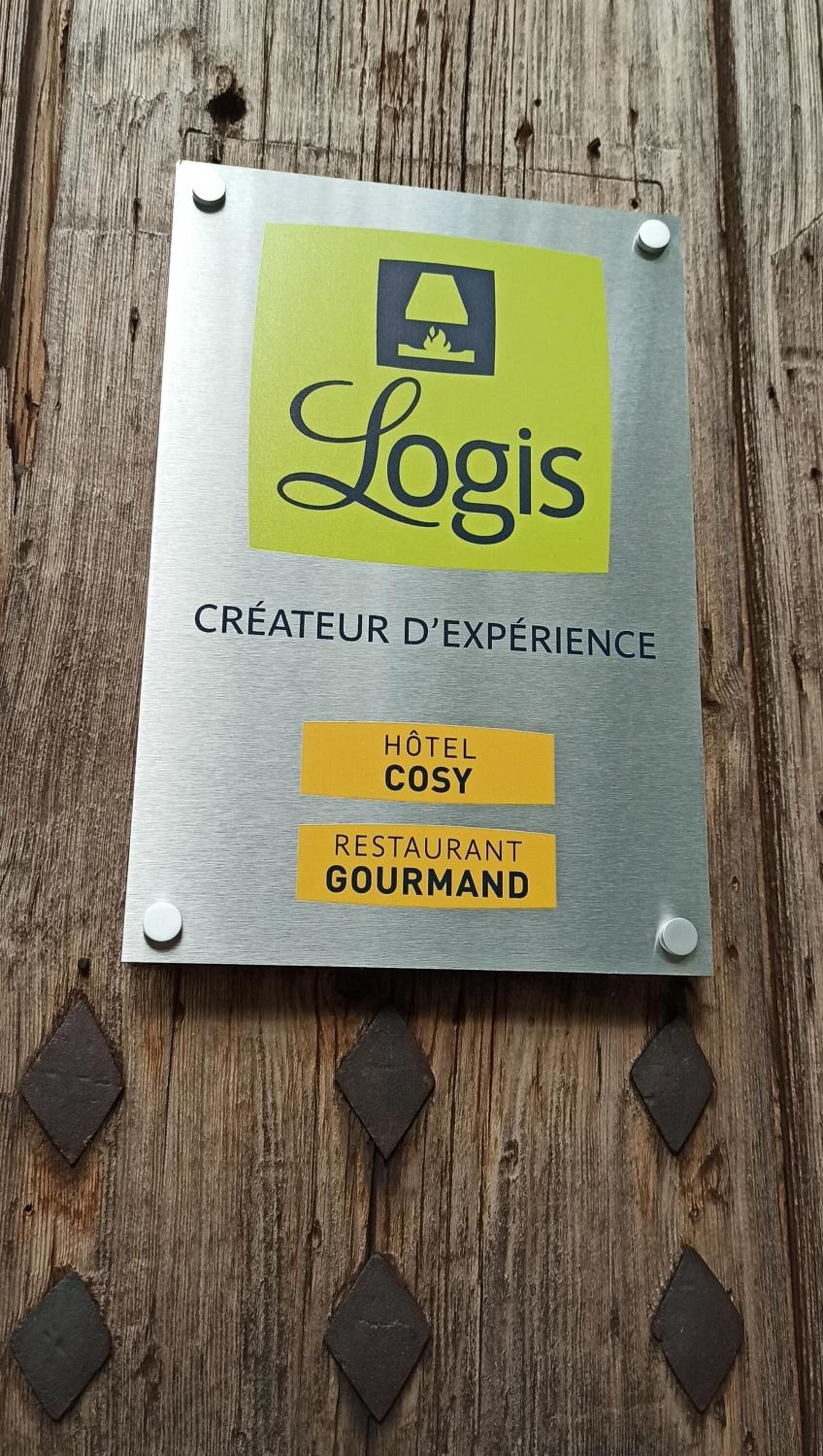 Eine Metallplakette mit einem Logo und Text, der angibt, dass es sich um einen Anbieter von Erlebnissen handelt, verbunden mit Hotel Cosy und Restaurant Gourmand.