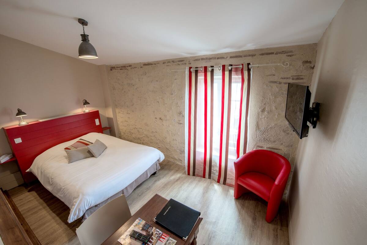 A bedroom with a red headboard, a single bed, a red chair, a wooden coffee table with books, a TV mounted on the wall, and a window with striped curtains.