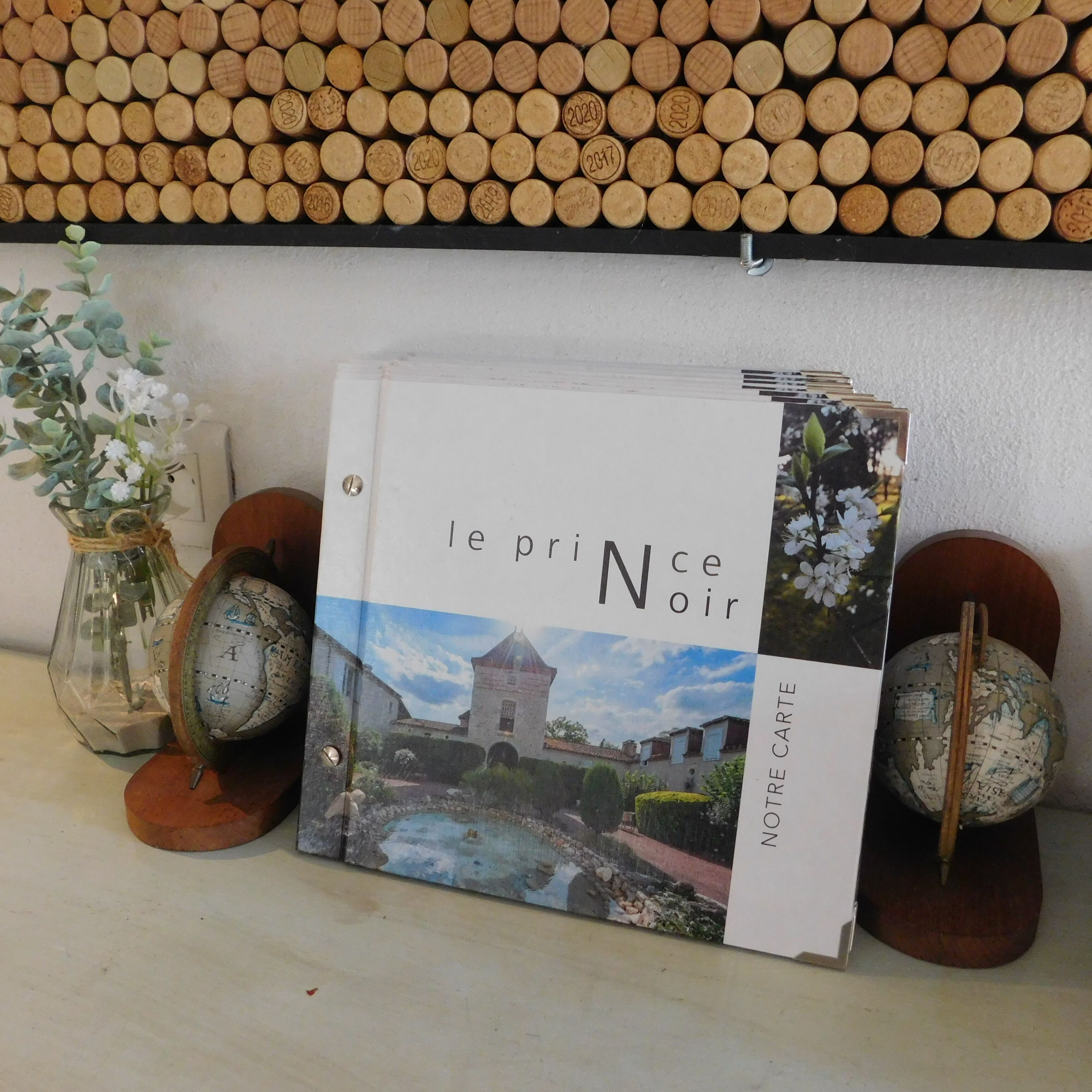 Un libro titulado 'le prince noir' con una imagen de portada que muestra un paisaje, ubicado en un estante junto con tapones de corcho y objetos decorativos.