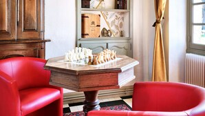 Una habitación con un tablero de ajedrez, sillas rojas, un aparador de madera y una estantería con diversos objetos.