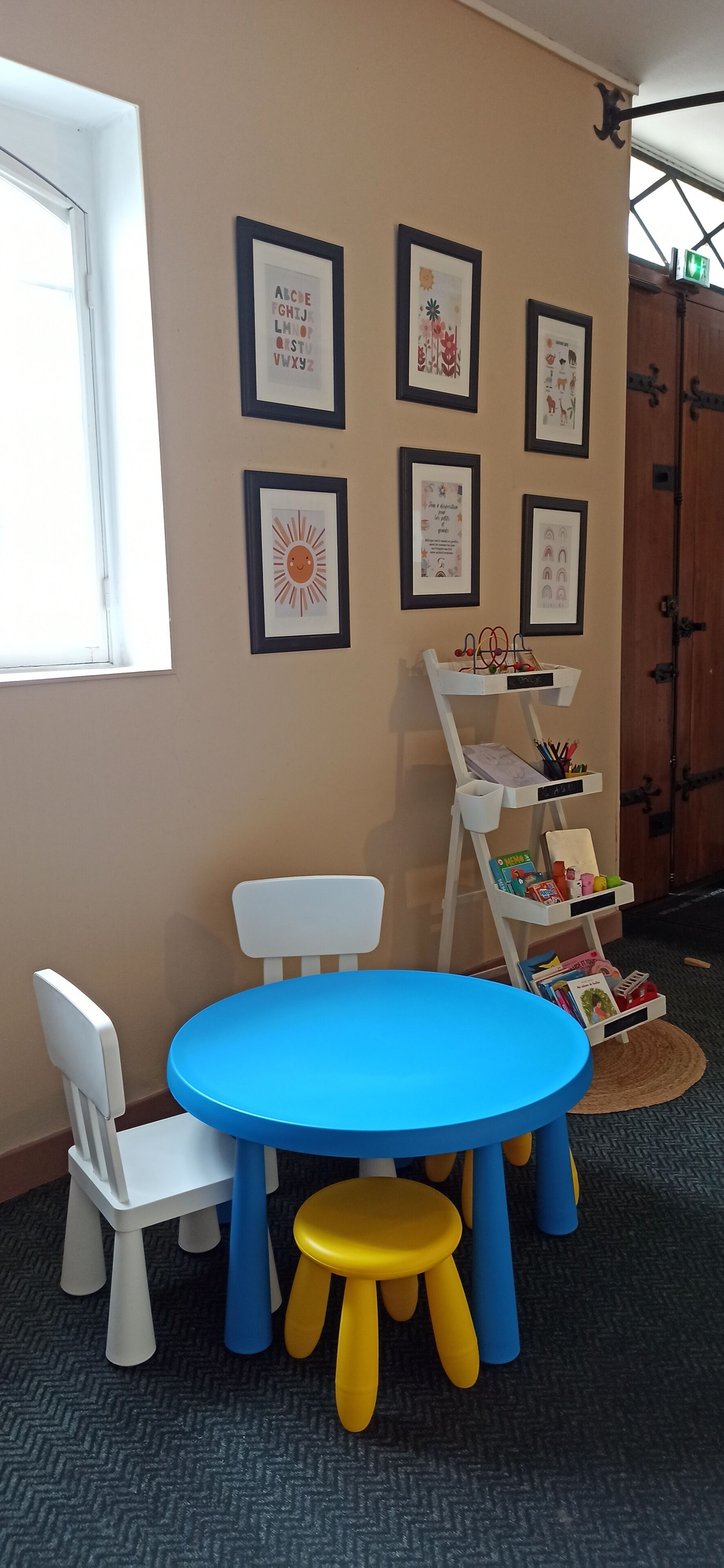 A children's play area with a blue table, white chairs, and yellow stools. There are framed pictures on the wall and a white shelf with books and toys.