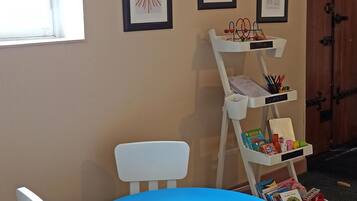 A children's play area with a blue table, white chairs, and yellow stools. There are framed pictures on the wall and a white shelf with books and toys.