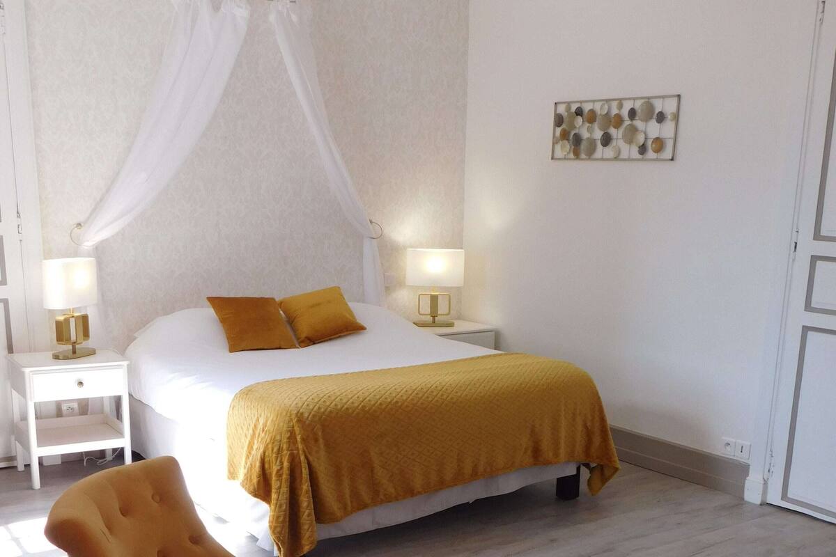 A bedroom with a bed, two bedside tables, a nightstand, a chair, a round table, and a wall art piece.